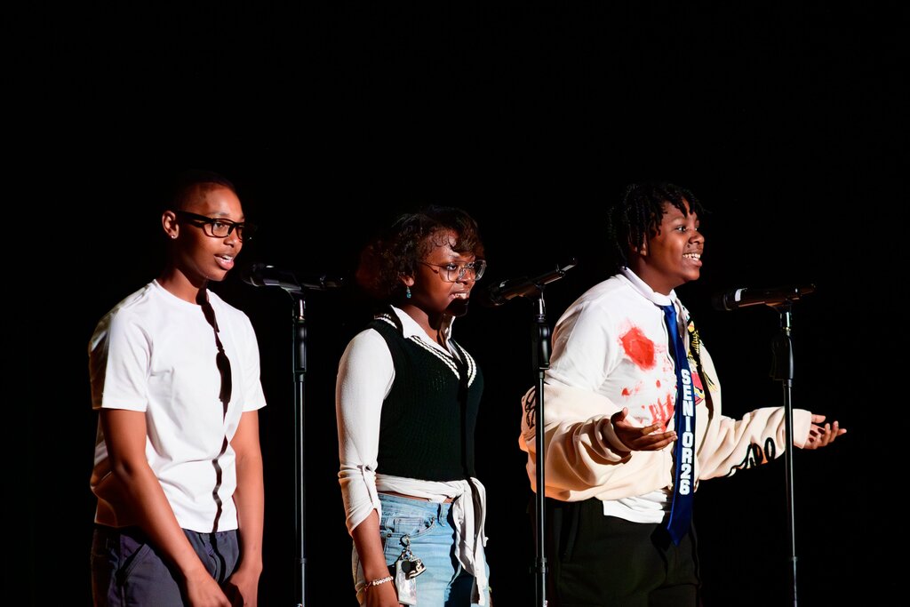 Three young performers stand in front of microphones on a dark stage, delivering a spoken word or poetry performance.