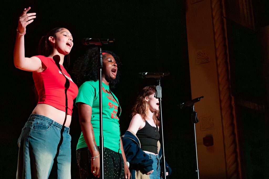 Three young women stand on stage in front of microphones, performing or speaking under warm stage lighting.