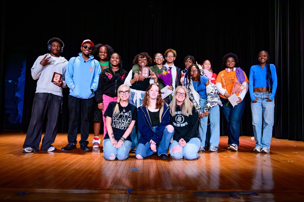 Group of diverse young people posing on stage, some holding awards, smiling under bright lights with a dark curtain backdrop.