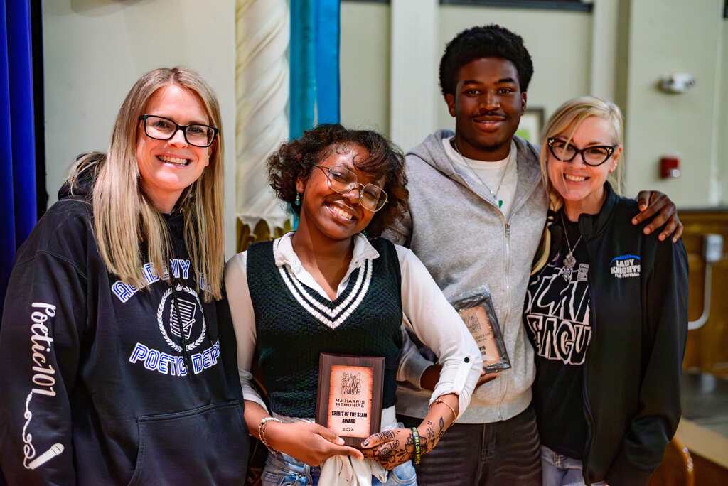 Four diverse young people standing together indoors, two holding plaques, dressed casually in hoodies and sweaters.