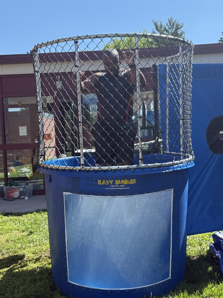 Man inside a blue dunk tank with safety netting, preparing to be dunked outdoors on a sunny day.