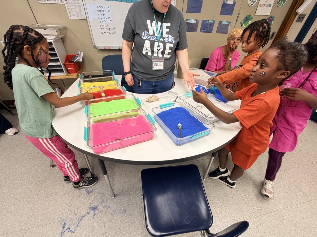 Children engaging in a colorful sensory activity with trays of sand while an adult guides them around a round table in a classroom.