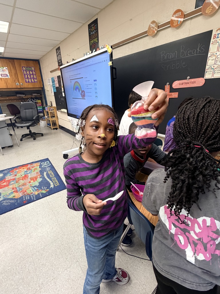 Child in a purple striped shirt holding a colorful layered sand art bottle in a classroom setting.