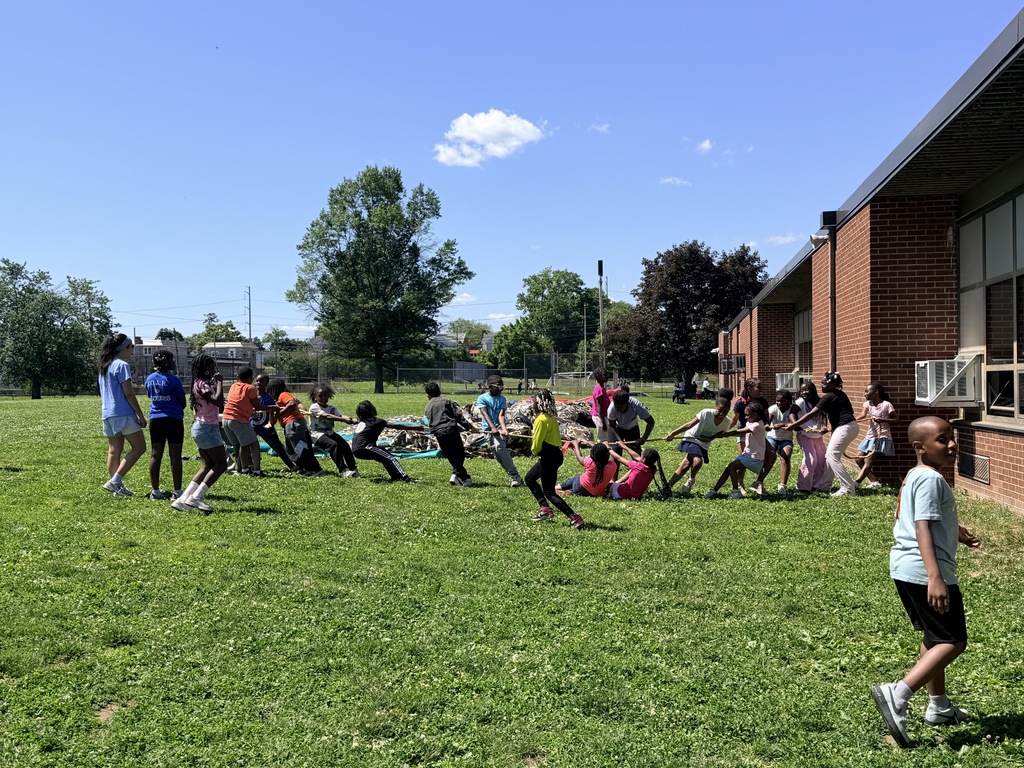 Children playing tug-of-war on a sunny grassy field near a brick building under a clear blue sky.