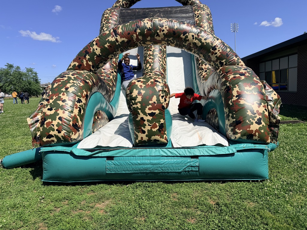 Two children sliding down a large camouflage-patterned inflatable slide on a sunny day outdoors.
