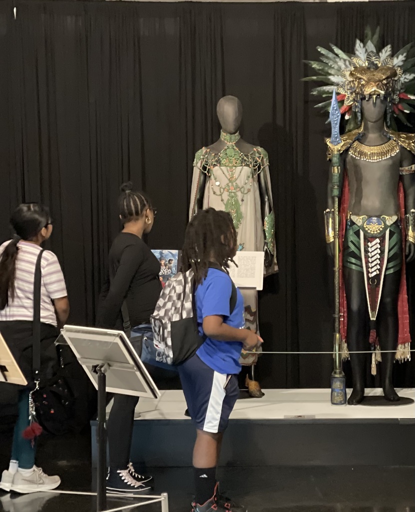Students view detailed fantasy and Afrofuturist costumes displayed on mannequins, including an ornate warrior-inspired outfit with a ceremonial staff.