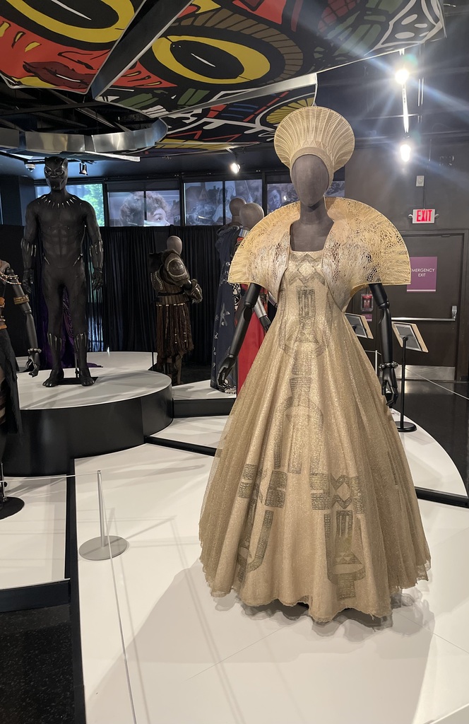 Gold gown with a dramatic sculptural collar and headdress displayed among costumes from film and cultural history exhibits.