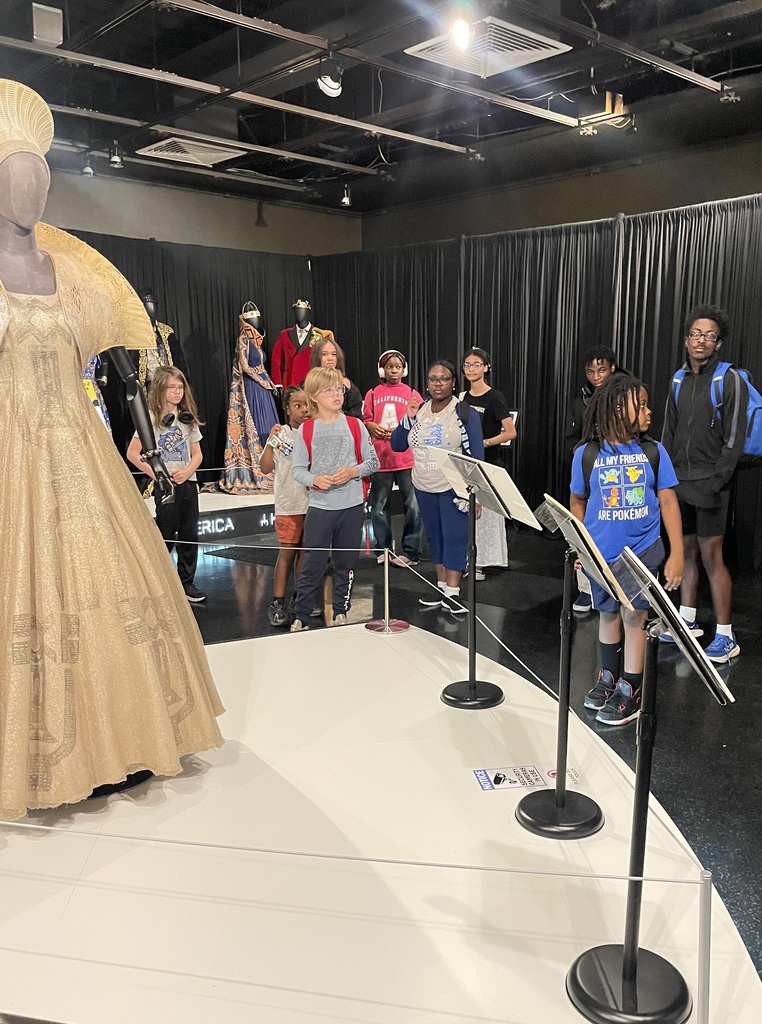 Students explore a costume exhibit featuring elaborate gowns and historical garments displayed on mannequins in a museum gallery.