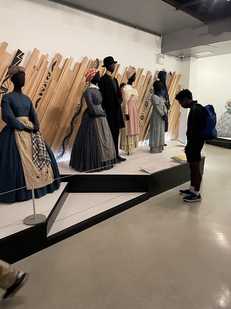 Student studies nineteenth-century clothing displayed on mannequins, including dresses, aprons, and formal attire in a museum exhibit.