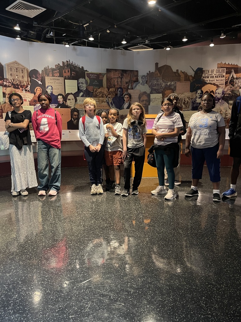 Students stand in a museum gallery listening to a presentation in front of a large exhibit featuring historical figures, documents, and artwork.