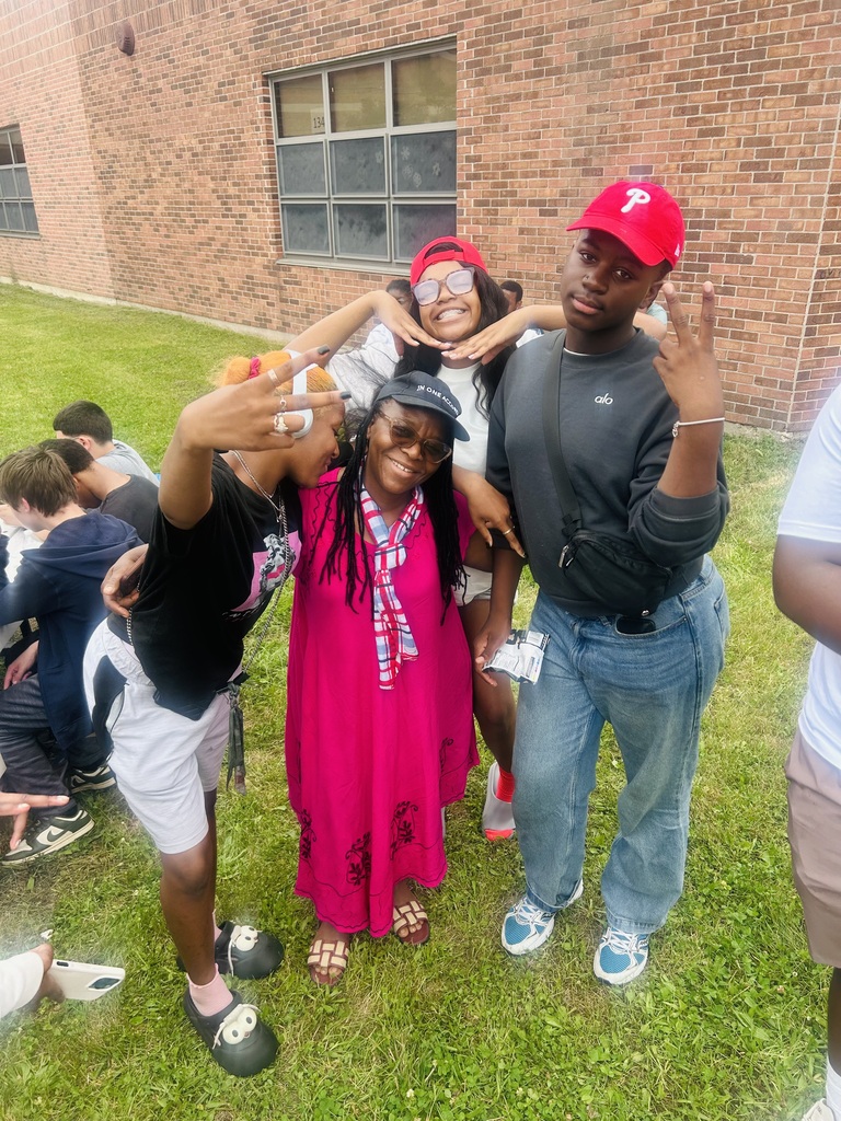 Group of smiling students poses outdoors on a school lawn with a staff member in a bright pink dress, making playful hand gestures in front of a brick building.