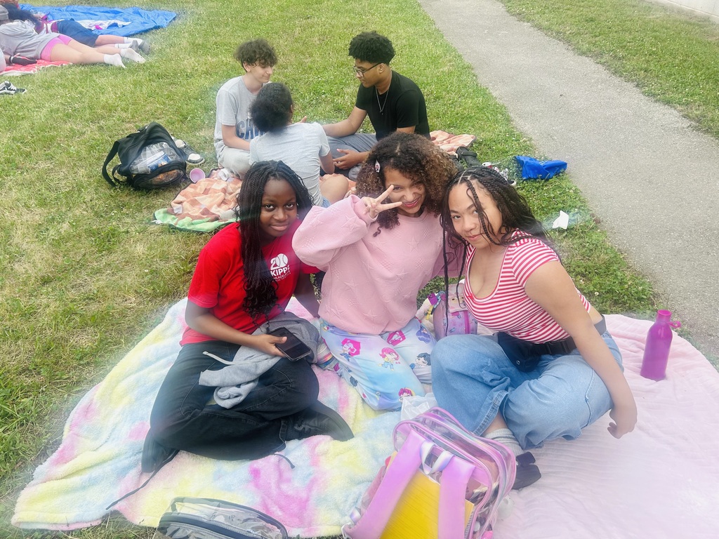 Three students sit together on a blanket during an outdoor school gathering, smiling and posing for the camera while classmates relax on the grass nearby.