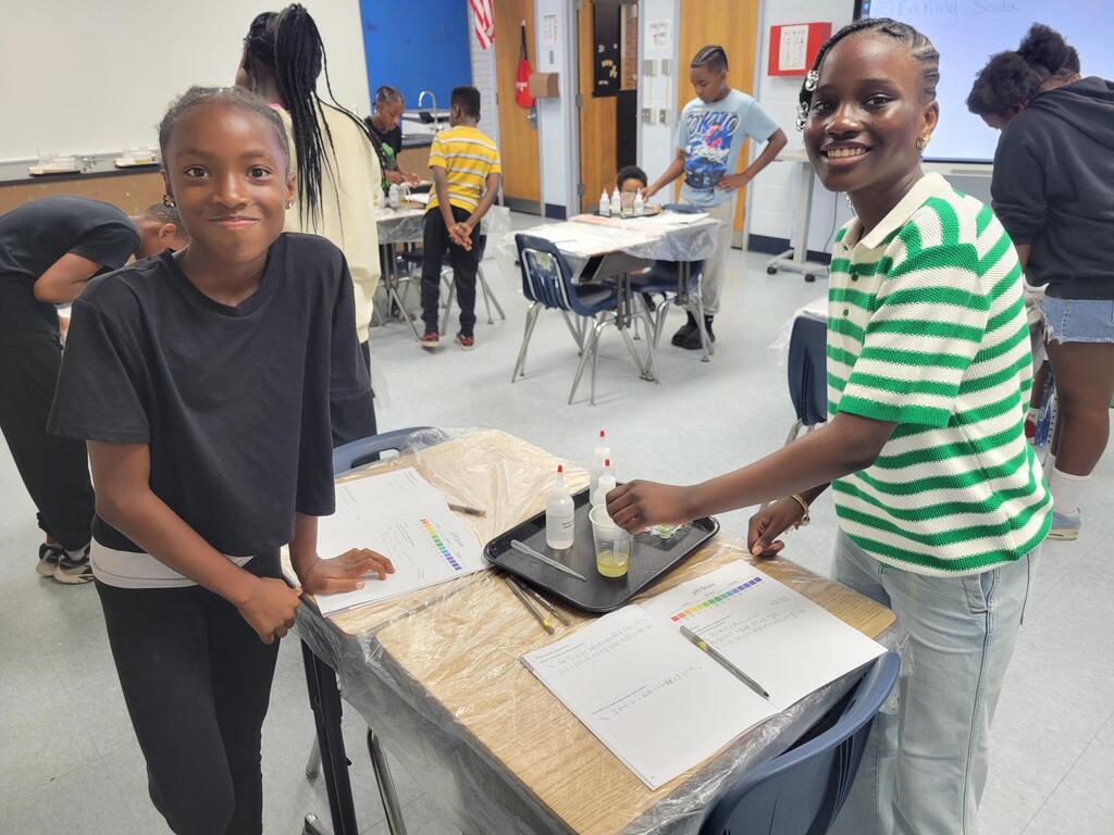 Two smiling students conduct a science experiment with liquids at a classroom table covered in plastic.