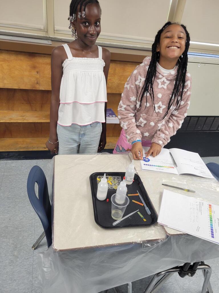 Two young girls stand by a classroom table with science experiment materials and open notebooks, smiling and engaged.