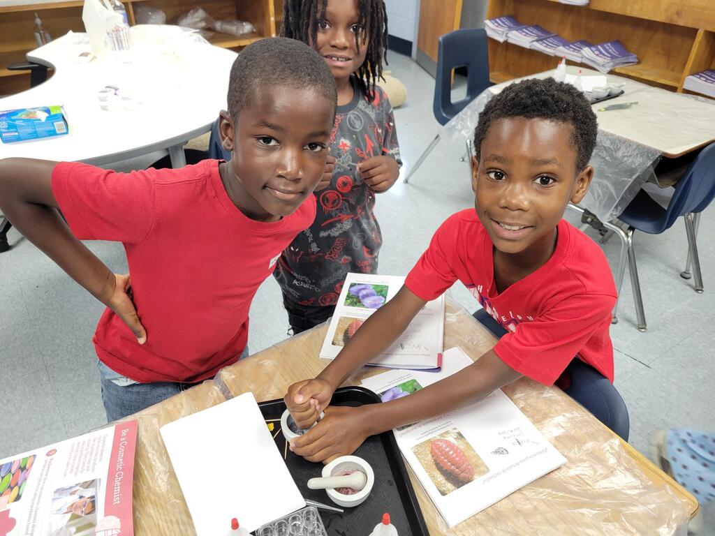 Three children engaged in a hands-on science activity with corn samples and educational materials on a classroom table.