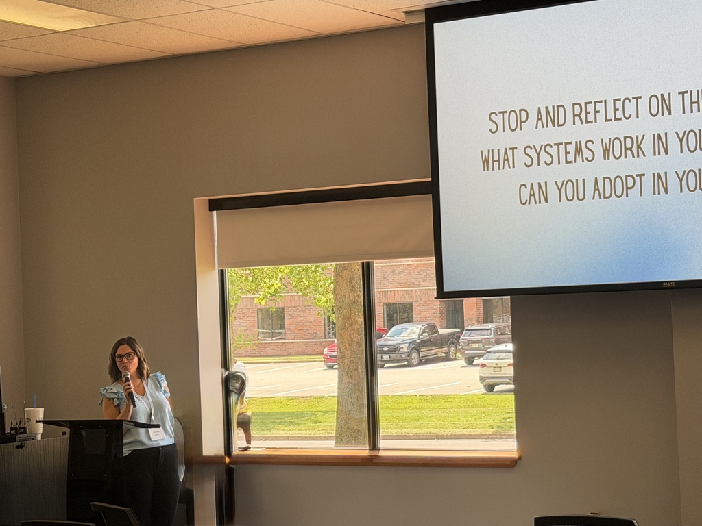 Presenter speaks at the front of a conference room beside a presentation encouraging participants to reflect on systems that support success.