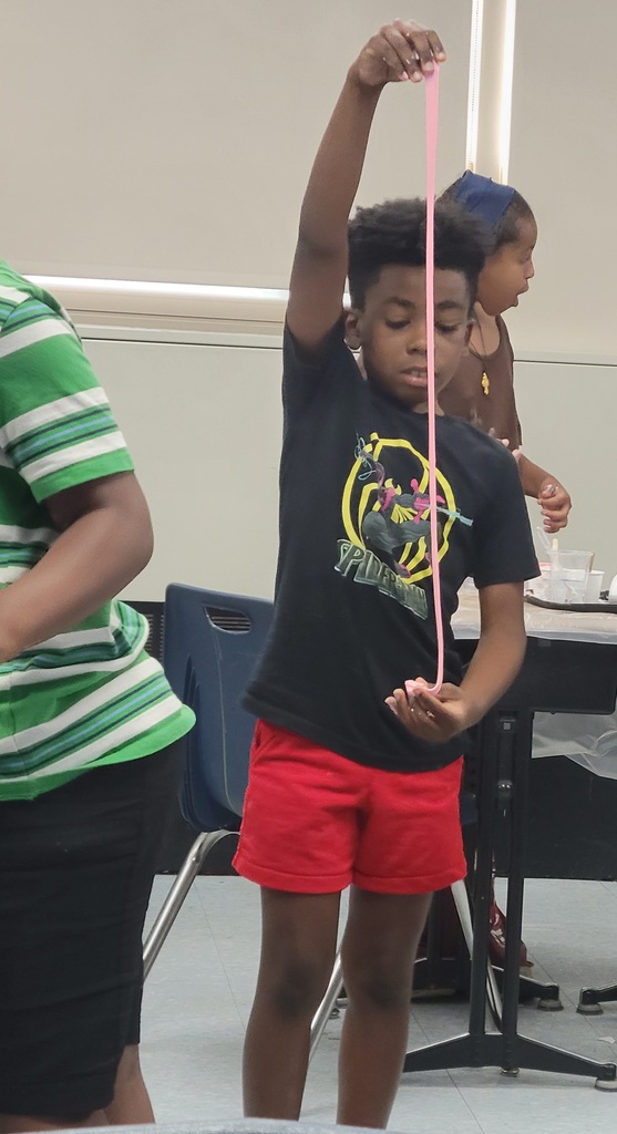 Boy in a black Spider-Man shirt stretching pink slime vertically in a classroom setting.