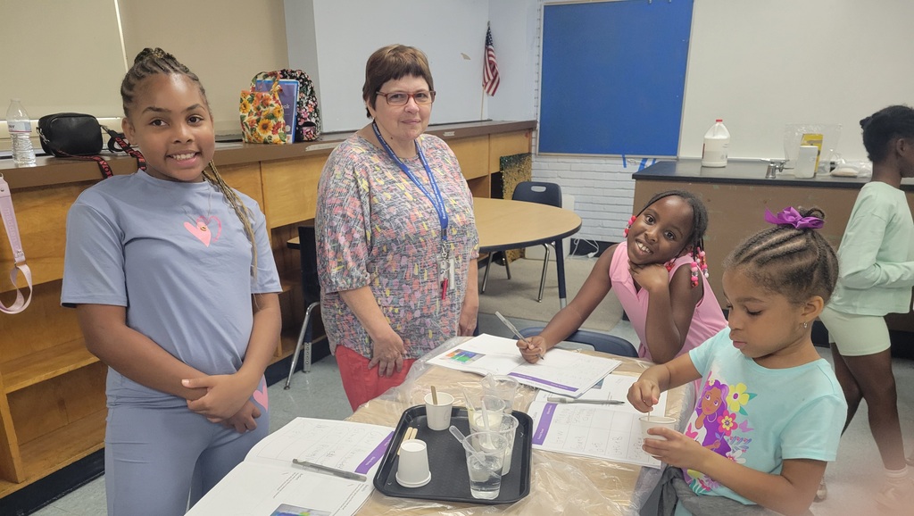 A teacher stands with three young girls engaged in a classroom science experiment involving liquids and measuring cups.