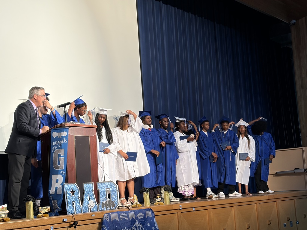Graduates in blue and white caps and gowns stand onstage holding diplomas and adjusting their tassels during a ceremony.
