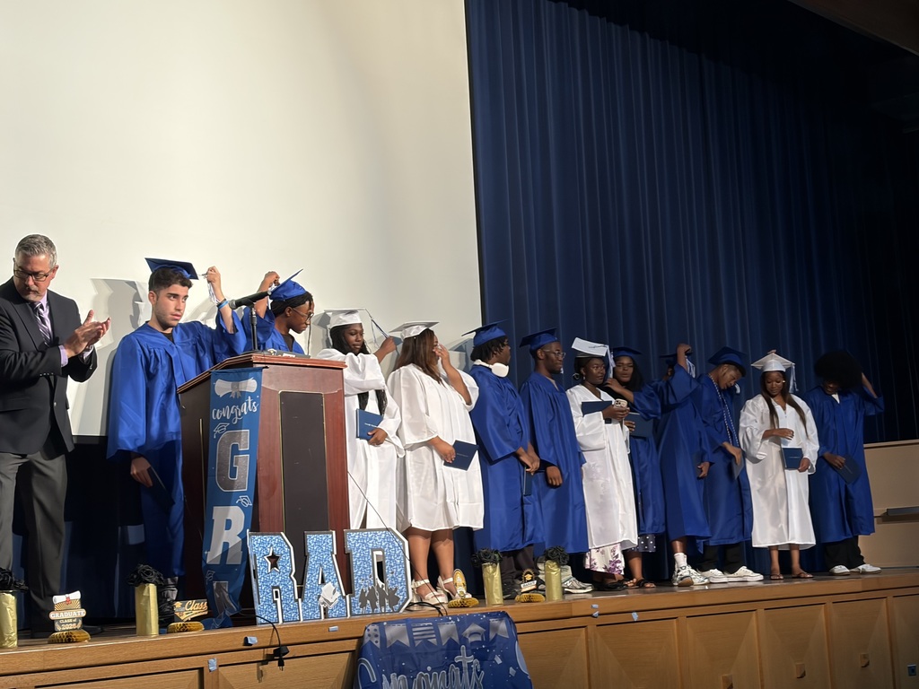Graduates in blue and white caps and gowns stand onstage holding diplomas and adjusting their tassels during a ceremony.