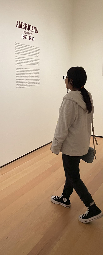 Person with glasses and a ponytail reading a museum wall text titled 'AMERICANA 1850-1950' in a minimalist gallery space.