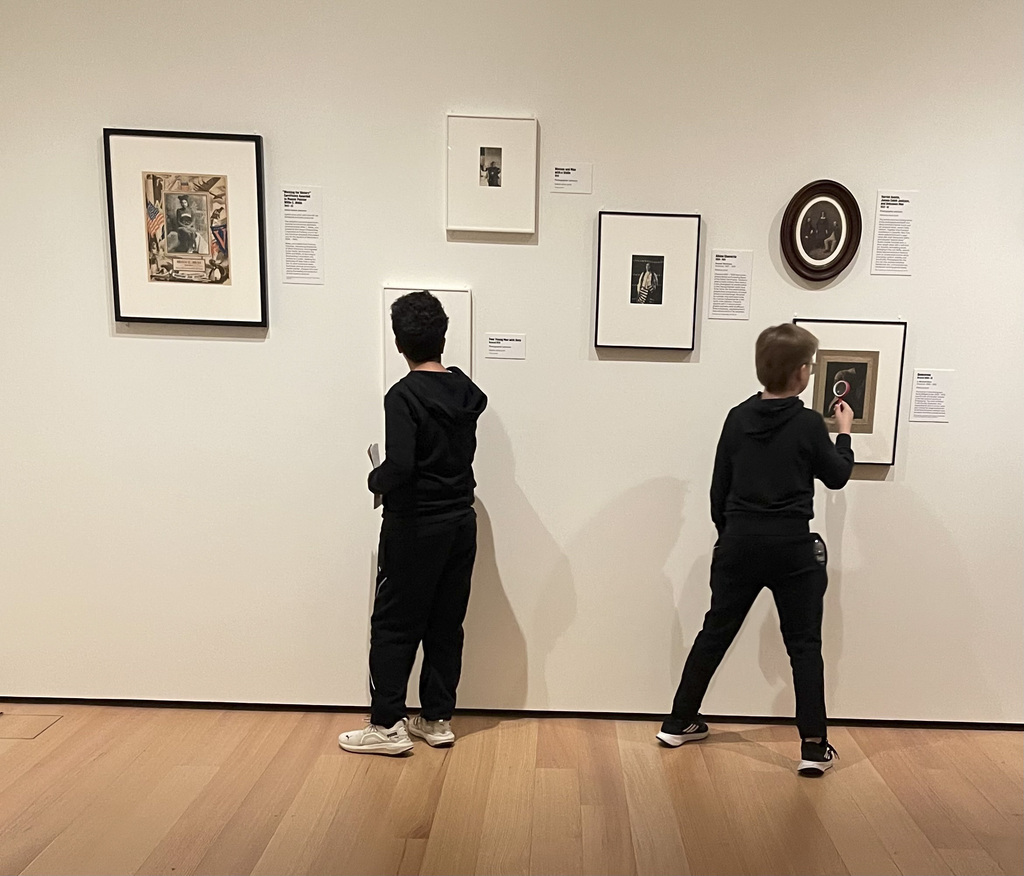 Two boys dressed in black closely examine framed artworks displayed on a white gallery wall with wooden flooring beneath.