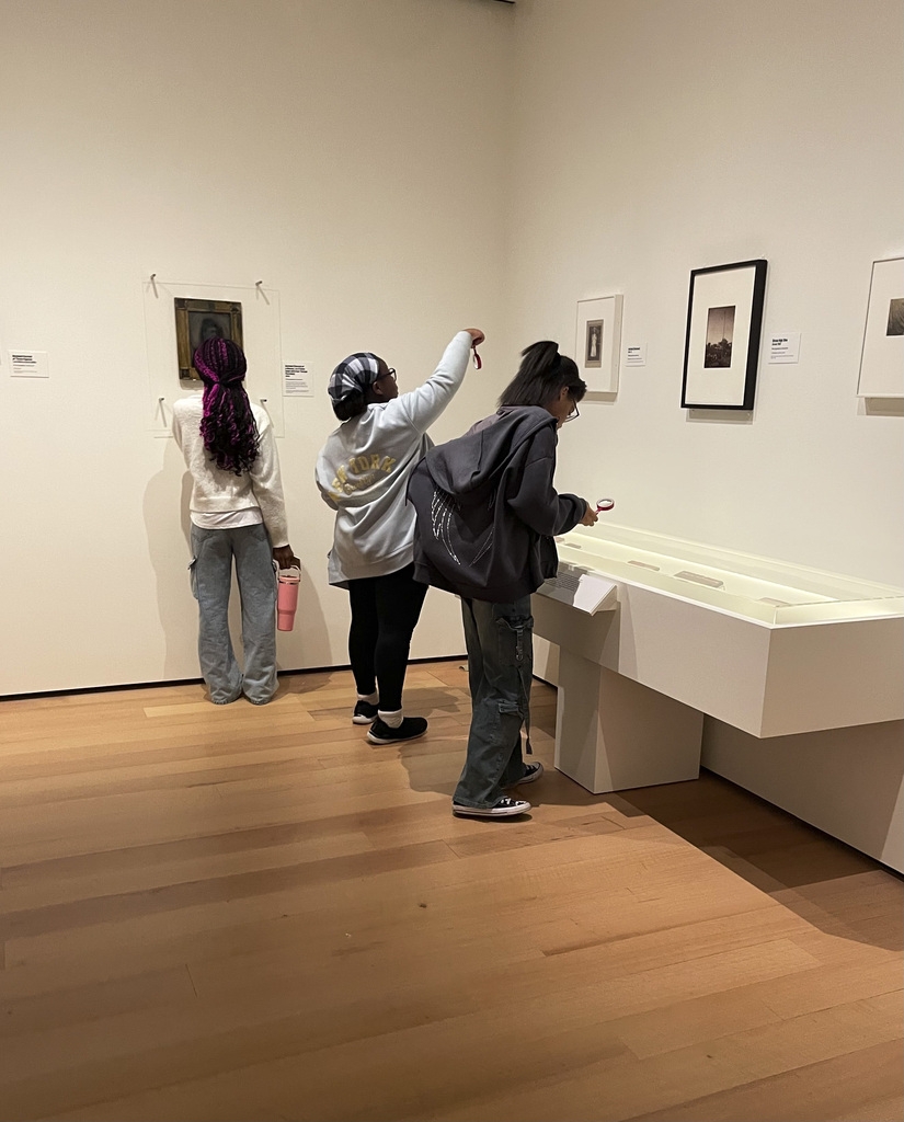 Three visitors closely examine framed artworks and display cases in a modern art gallery with wooden floors and white walls.