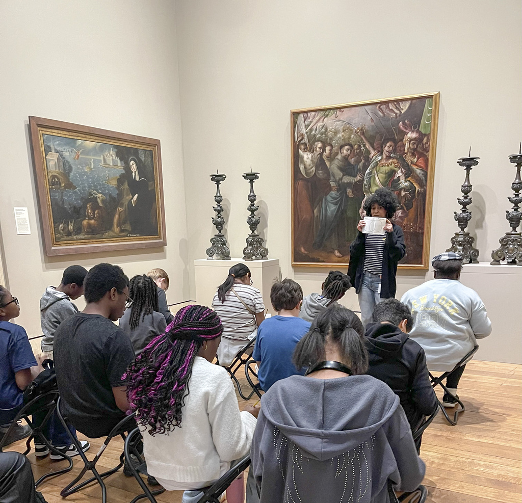A group of students seated on stools attentively listening to a guide in an art gallery with large classical paintings and ornate candlesticks on display.