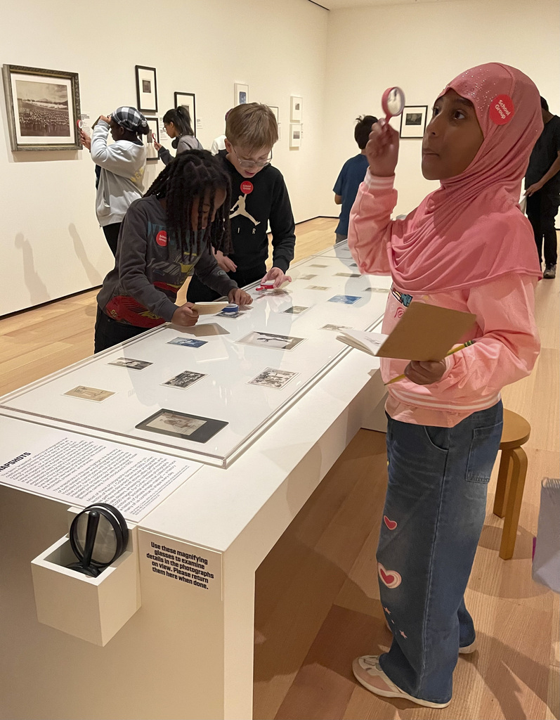 Children exploring historical photographs and documents on a museum display table, using magnifying glasses and taking notes.