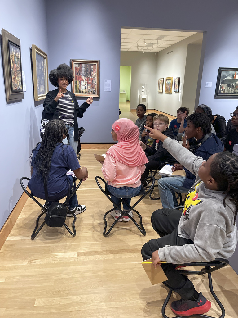 A museum guide enthusiastically explains artwork to a diverse group of children seated on folding chairs in a gallery.