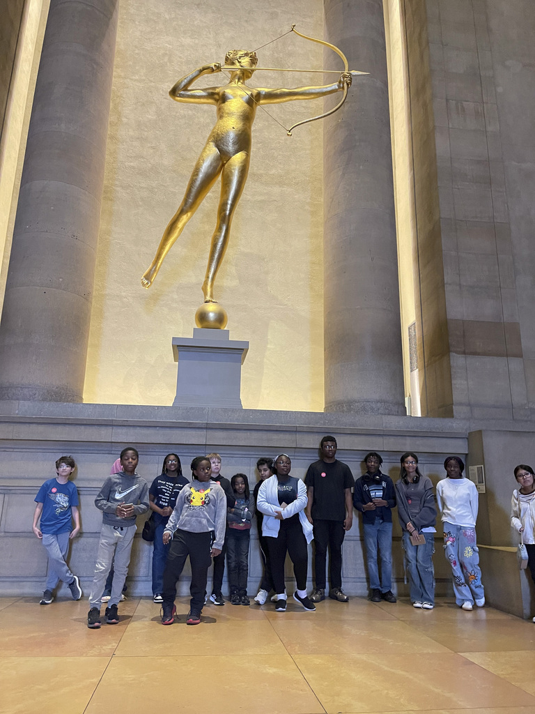 Group of diverse young people standing in front of a large golden statue of a female archer inside a grand hall.
