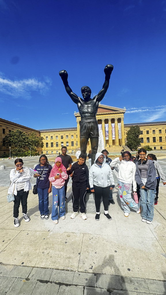 Group of children posing in front of the Rocky Balboa statue with the Philadelphia Museum of Art in the background under a clear blue sky.
