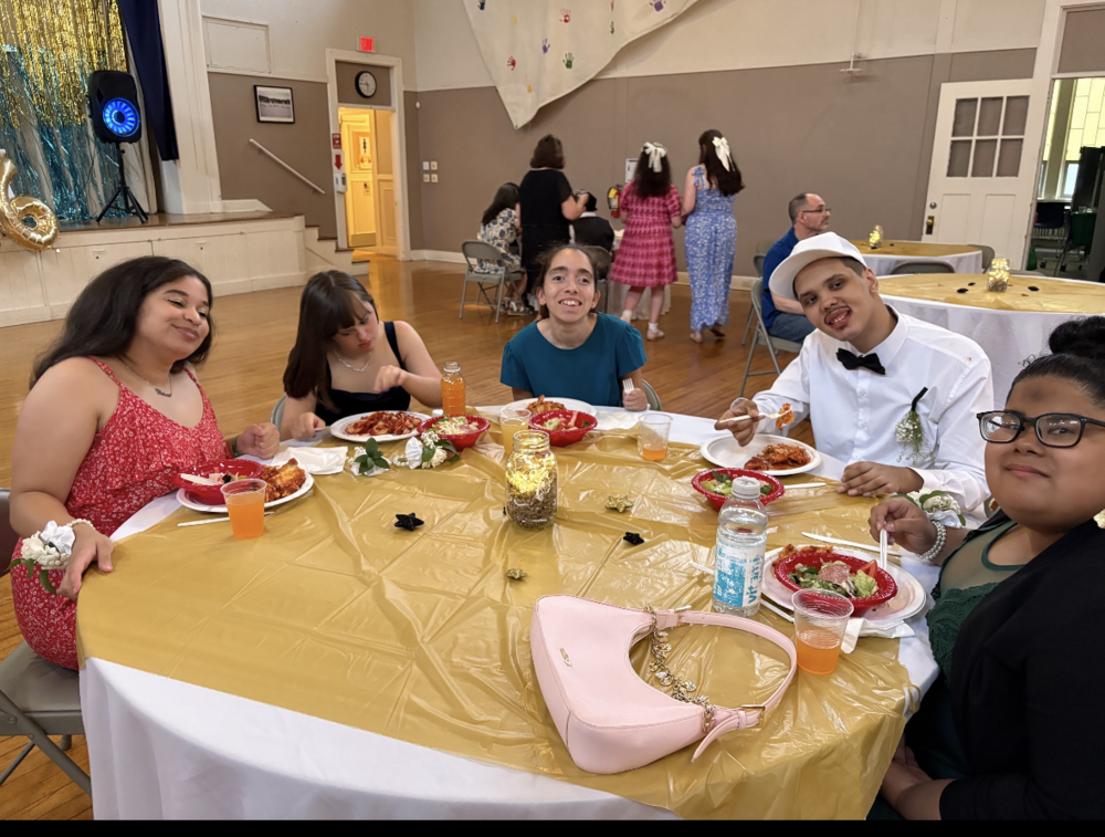 Five people are seated around a round table at a banquet or celebration in a community hall, smiling toward the camera while eating pasta and salad. The table is covered with a gold tablecloth and decorated with a lit mason jar centerpiece, white flowers, and small gold accents. A light pink handbag sits in the foreground. One person is dressed in a white shirt, black bow tie, and white cap with a boutonniere, suggesting a special occasion. In the background, other guests mingle and sit at tables, with a small stage featuring gold and blue decorations, a large gold number "6" balloon, and colorful handprint artwork hanging on the wall. The atmosphere is festive and welcoming.