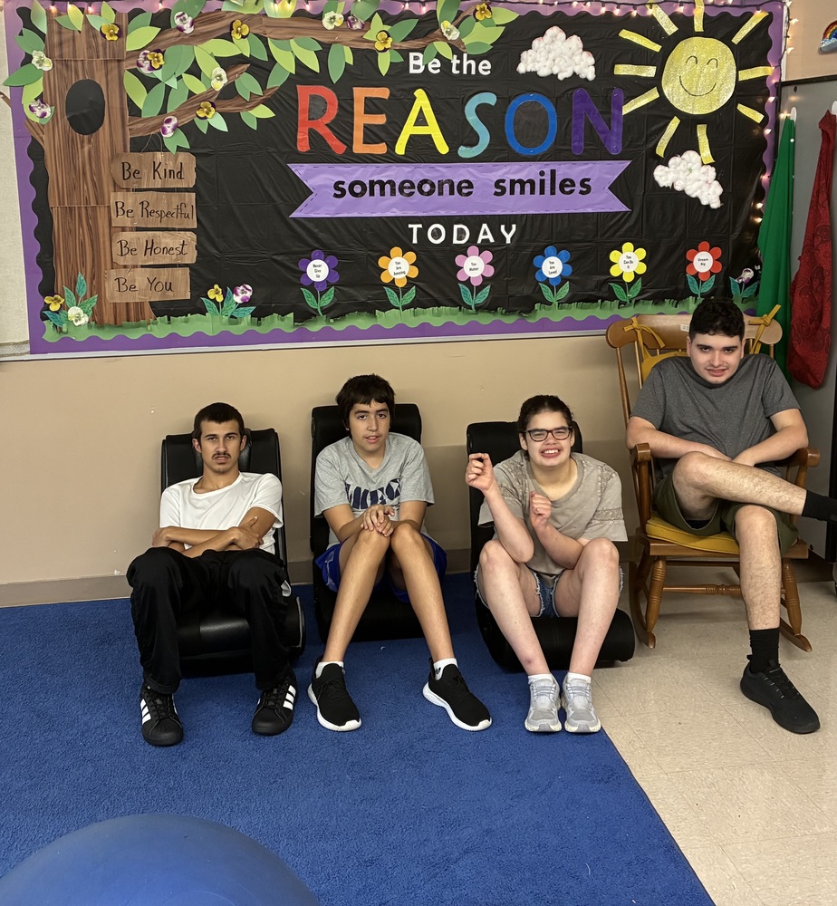 Four students seated in a SMEC classroom in front of a bulletin board that reads, "Be the REASON someone smiles TODAY." On the far left, a male student in a white t-shirt and black pants sits with arms crossed in a floor rocker chair. Beside him, a male student in a gray "SMEC" t-shirt and blue shorts sits smiling. To his right, a female student with glasses and a gray t-shirt smiles widely at the camera. On the far right, a male student in a dark gray t-shirt and shorts sits in a wooden rocking chair.