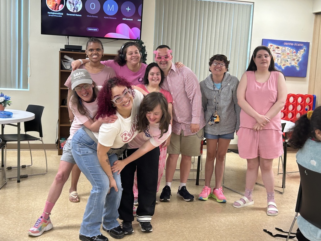 n the center foreground, three women are playfully posed together. One woman, wearing a white graphic t-shirt and blue jeans, leans forward with a wide grin. Another woman in a pink shirt and black pants leans against her, and a third woman in a grey "SBEC" hat and light blue shirt is partially crouched beside them.BackgroundBehind them, five more people stand in a row:On the left: Two women, one in a pink shirt and another in a maroon shirt, are smiling broadly.In the center: A man in a pink checkered button-down shirt and pink novelty glasses has his arms around the women next to him.On the right: A woman in a grey zip-up hoodie and denim shorts stands next to a woman in a matching pink sleeveless top and shorts set.