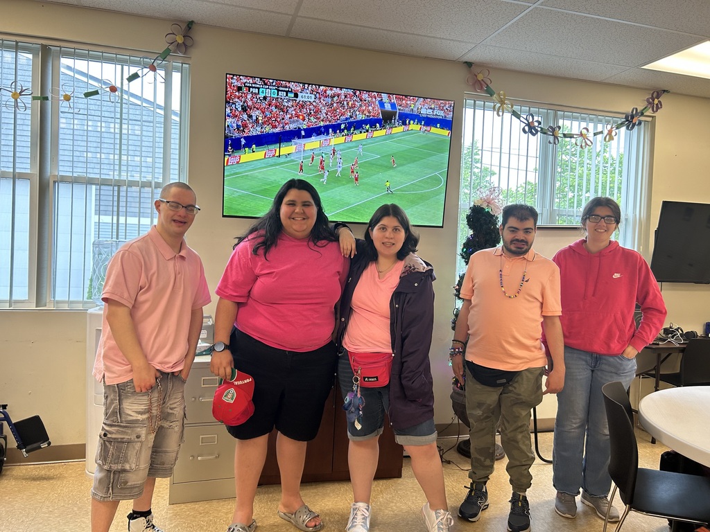 The image depicts a diverse group of five young adults standing together in what appears to be a brightly lit recreational room, smiling for a group portrait.