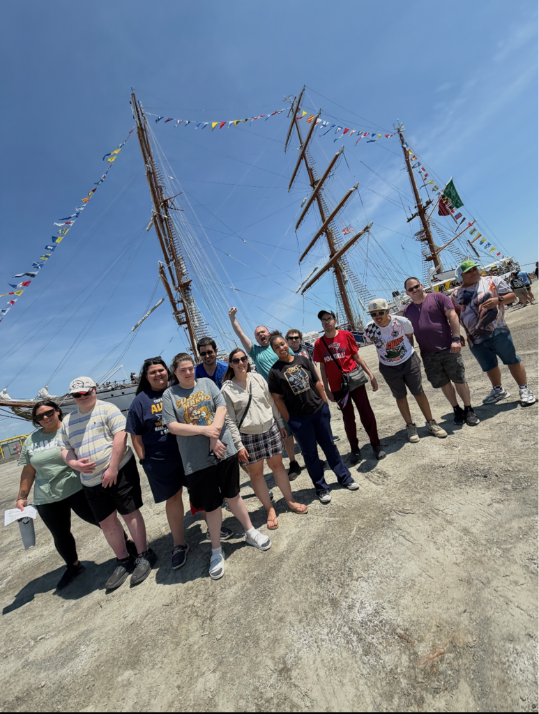 A group of eleven adults poses together outside on a sunny day in front of a large, three-masted tall ship decorated with colorful nautical flags under a clear blue sky.