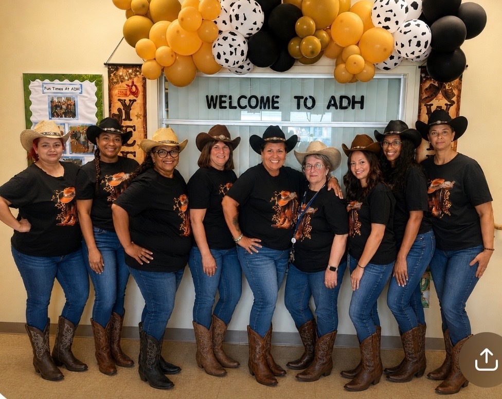 Eight women in black T-shirts, blue jeans, and cowboy boots pose together in front of a decorated “WELCOME TO ADH” display. They are wearing cowboy hats, with a large black, orange, and white balloon arrangement overhead. A bulletin board on the left reads “Fun Times At ADH.”