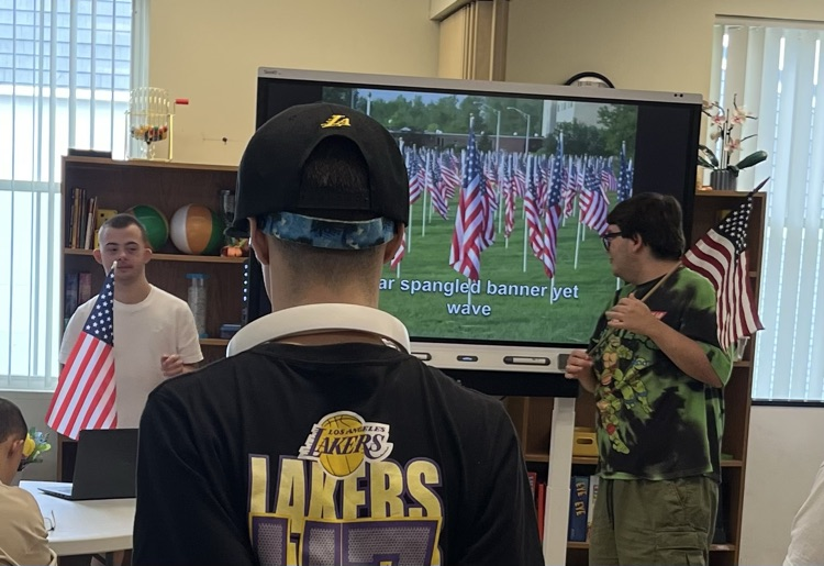 A room with young adults at tables watching a video on a large television