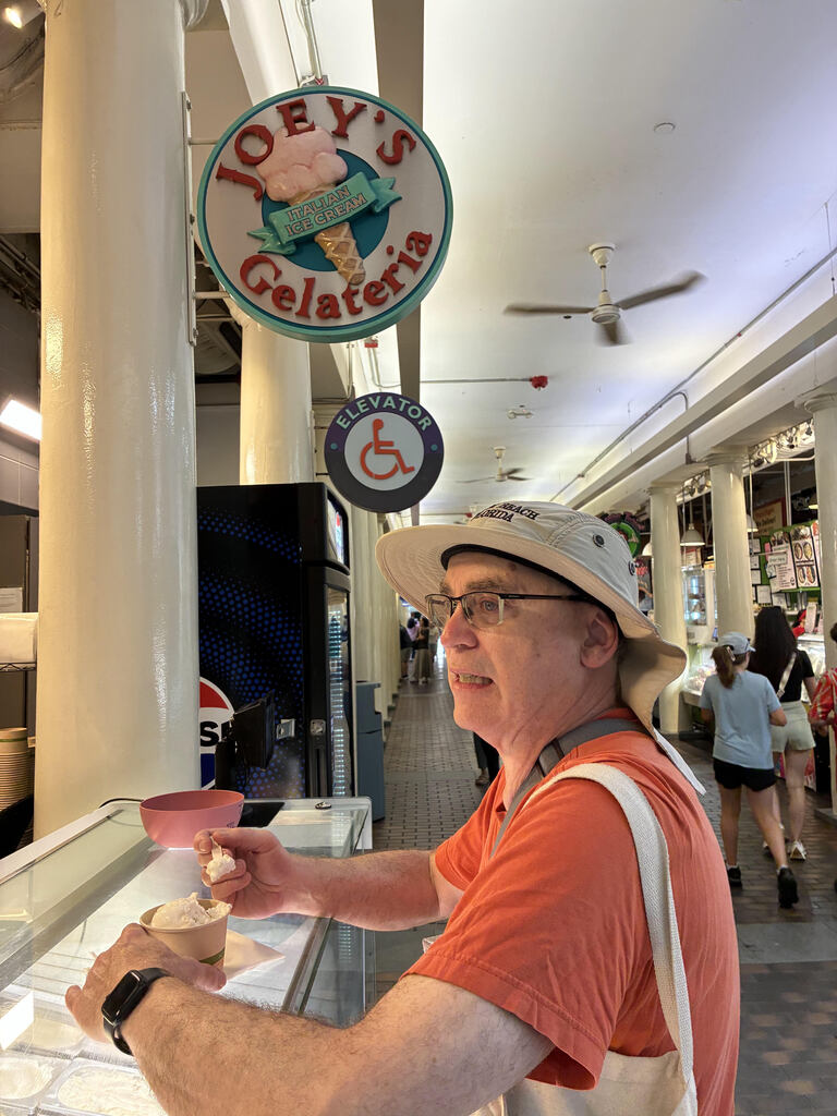 Man in orange shirt stands at ice cream counter eating cup of vanilla ice cream.