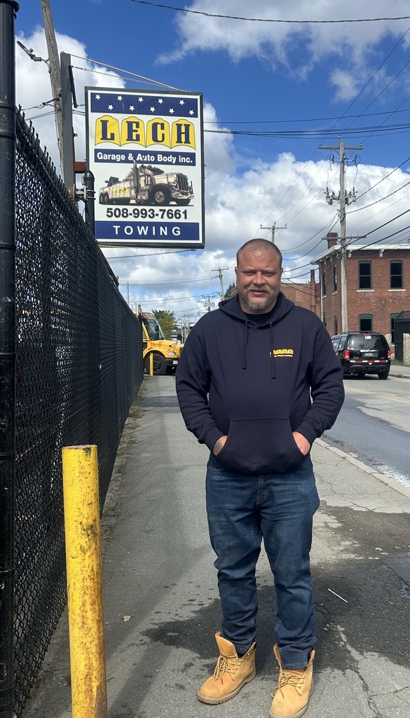Man in  work clothes stands on the sidewalk with auto body sign in the background.