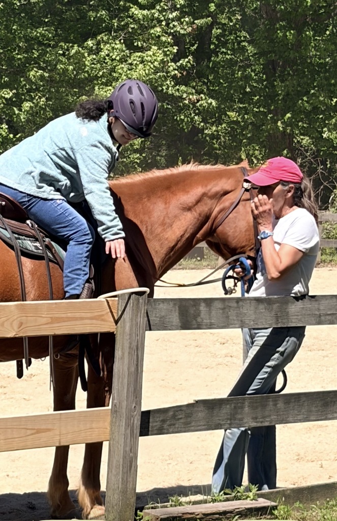 a Woman sitting on a horse picking up a ring off of a fence post with handlers alongside 