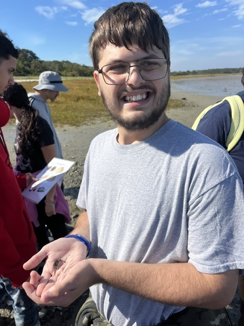 A student in a grey tshirt holding out a shell in his hand and other students are walking behind him