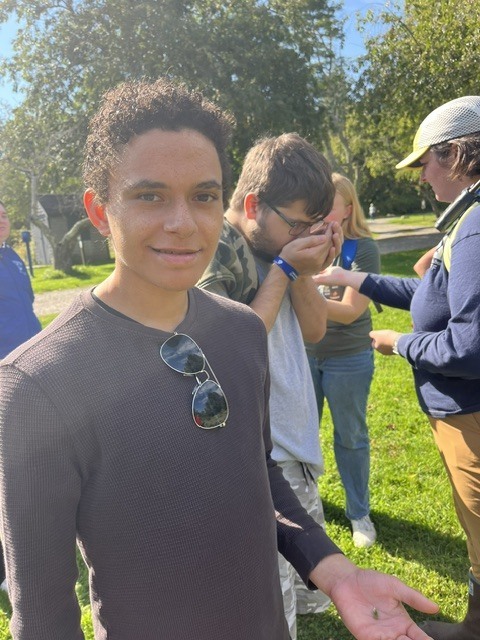 A student with sunglasses on his shirt with a small item from nature, another student is smelling something from nature