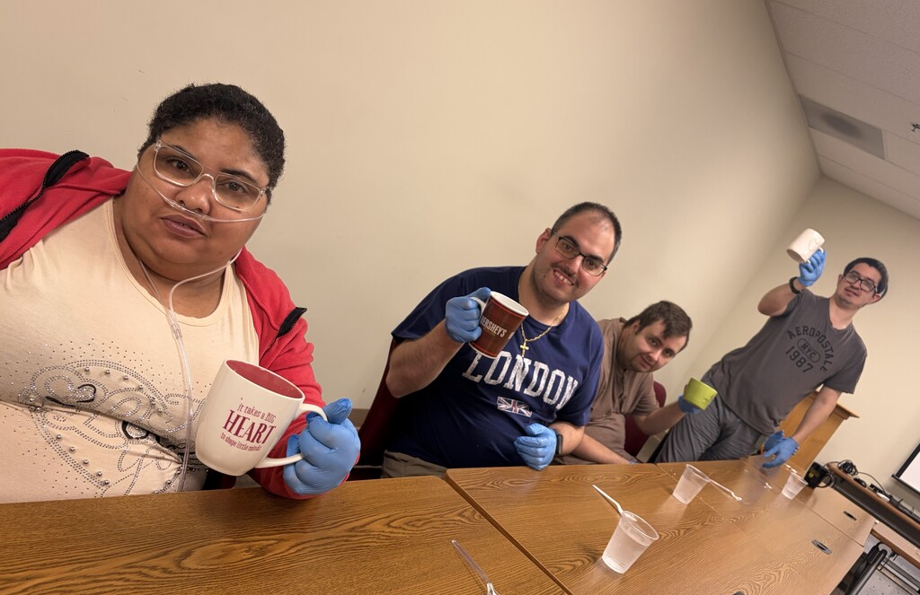 A woman and a group of me sit at a table with rubber gloves on holding up a coffee mug.