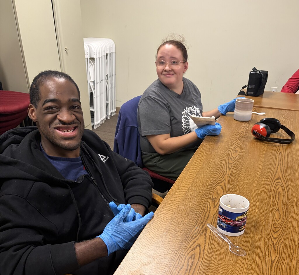 A man and a woman sit at a table wearing rubber gloves, with a mug and spoon on the table in front of them 