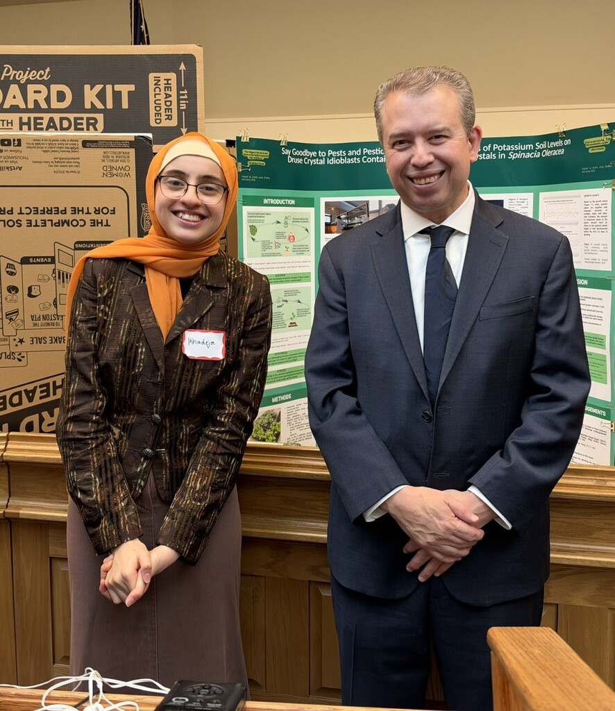 Southeastern student Khadeja Mukassabi smiles alongside Massachusetts Commissioner of Education Pedro Martinez at the State House, standing in front of her award-winning science project display on spinach, soil potassium levels, and pest management.