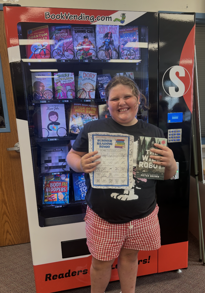 Smiling elementary school student standing in front of a red, black, and white Book Vending Machine filled with children’s books.