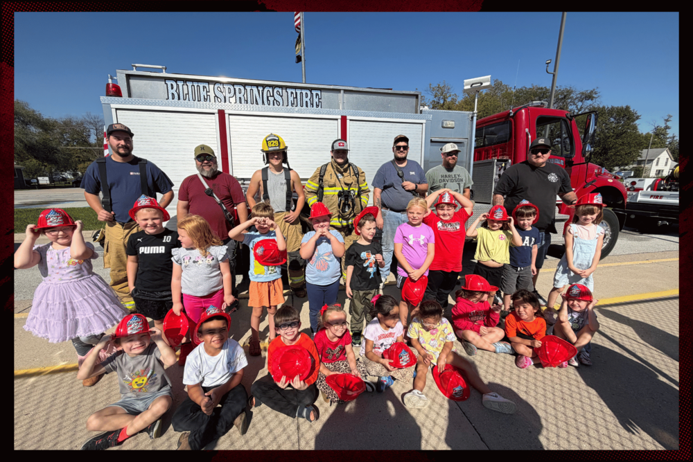 Kindergarten students pose for a group photo with members of the Blue Springs Fire Department in front of a fire truck during Fire Safety Week. Many students are wearing red firefighter hats as they celebrate a fun day of learning about fire safety and the important work firefighters do to keep our community safe.