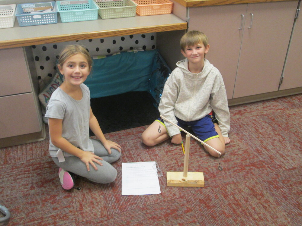 Two third-grade students sit on the classroom floor beside a student-built moving sculpture. The sculpture consists of a wooden base with a vertical wooden dowel and attached strings, rods, and hooks. A worksheet with instructions or notes rests on the floor nearby. The students are smiling and looking toward the camera. Classroom tables, storage cabinets, colorful bins, and a carpeted floor are visible in the background.